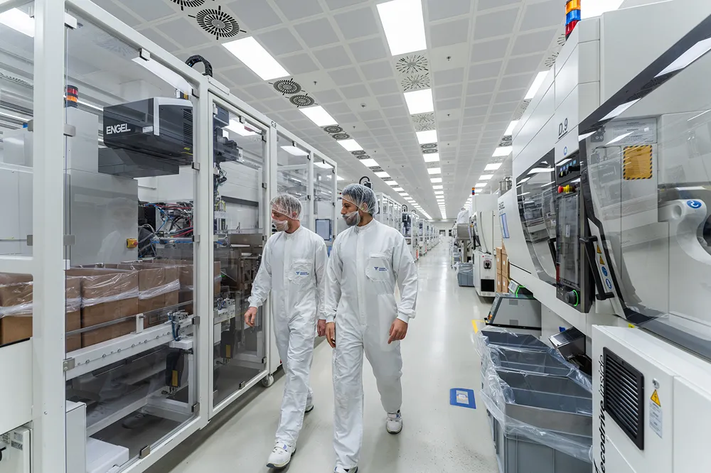 Two people in white cleanroom suits, hairnets, and face masks walk through a bright, modern manufacturing facility with large machines and equipment on both sides.