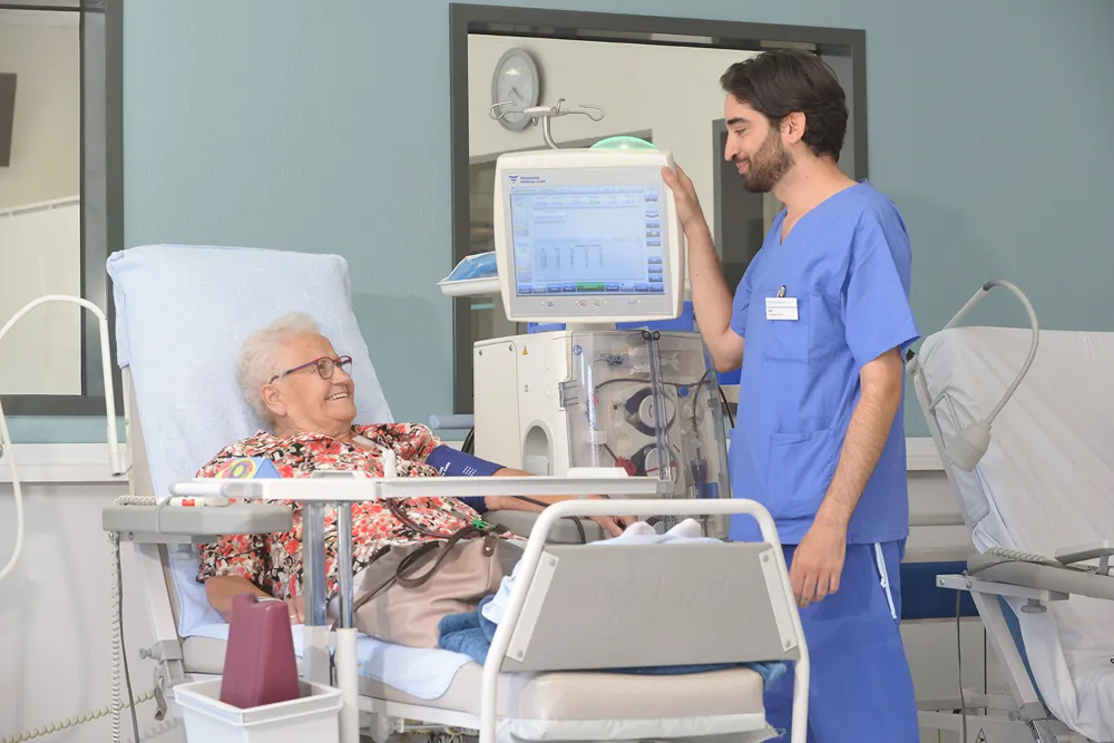 An elderly woman sits in a hospital bed, smiling at a healthcare worker in blue scrubs who stands beside her, adjusting medical equipment and monitoring her treatment.