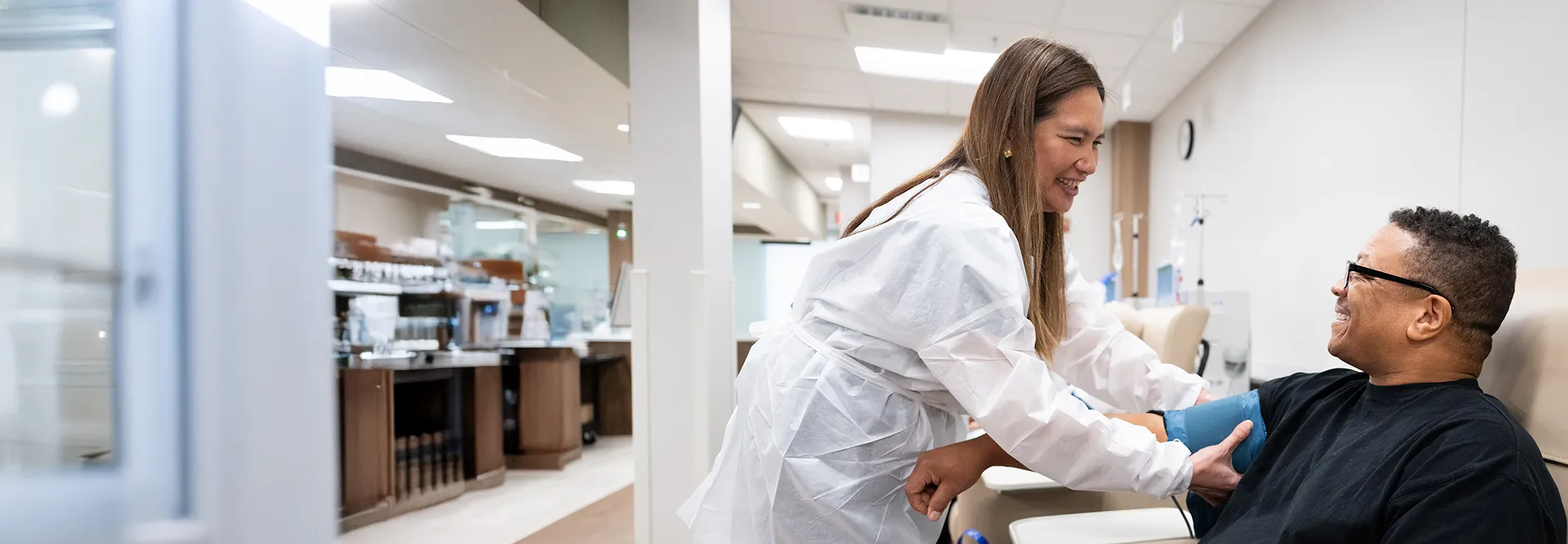 A healthcare worker in a white coat smiles while taking a blood pressure reading from a seated patient in a medical clinic. The patient smiles back, and the clinic is bright and modern.