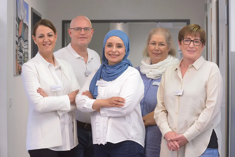 Five diverse professionals stand side by side indoors, smiling at the camera. They wear business casual attire and name badges, conveying a friendly, welcoming, and inclusive team environment.