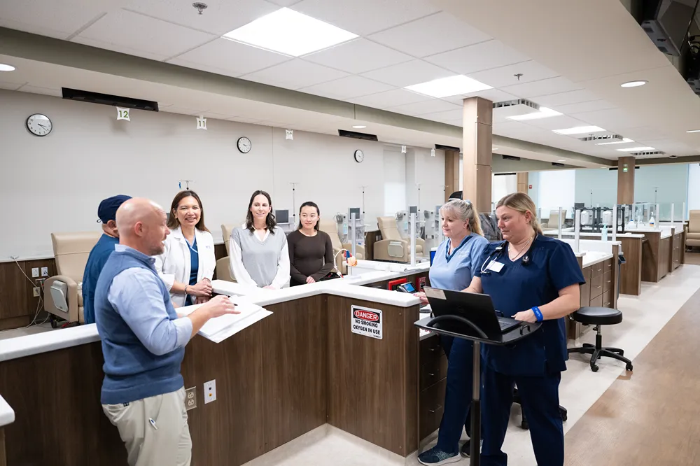 A group of healthcare professionals stand around a nurses’ station, discussing paperwork and using a laptop. The setting is a modern medical facility with bright lighting and clean, organized surroundings.