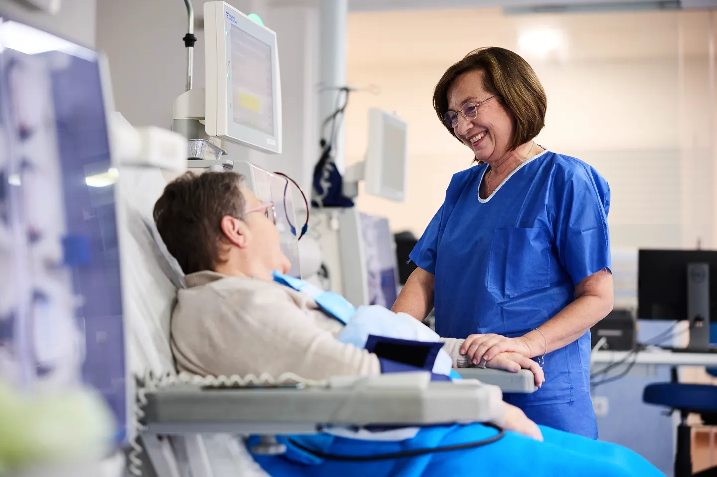 A nurse in blue scrubs smiles and holds the hand of a patient lying in a hospital bed, creating a caring and supportive atmosphere in a medical setting.