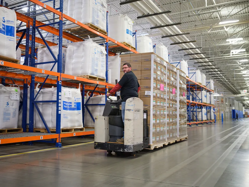 A worker drives a forklift carrying a large pallet of boxes through a warehouse with tall shelves filled with large white bags. Bright overhead lights illuminate the spacious, organized storage area.