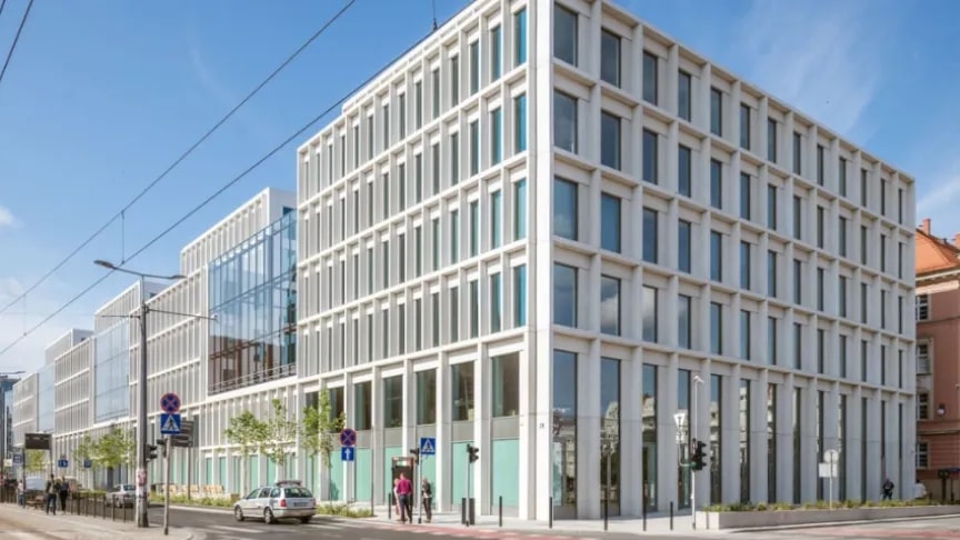 Modern office building with large glass windows and white grid-like framing, located on a city street with parked cars, street signs, and a few pedestrians on a sunny day.