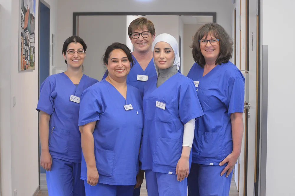 Five female healthcare professionals in blue scrubs stand together in a hallway, smiling at the camera. One woman wears a white hijab, and all have name badges, conveying a friendly medical team atmosphere.