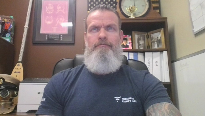 A man with a long gray beard sits in an office, wearing a navy blue T-shirt. Behind him are shelves with binders, books, trophies, framed photos, and a wall-mounted musical instrument.