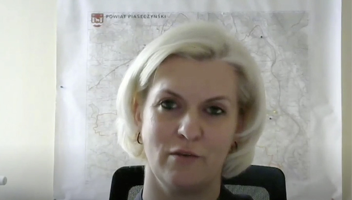 A woman with short blonde hair sits in front of a large map hanging on the wall, looking toward the camera.