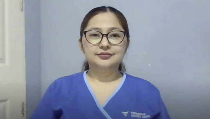 A woman with dark hair pulled back, wearing blue scrubs, sits indoors against a gray background, looking slightly to the side with a neutral expression.