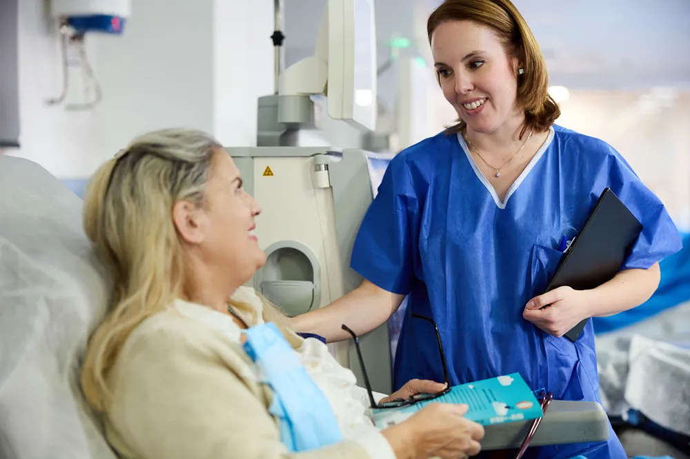 A nurse in blue scrubs smiles and speaks with a seated patient holding a brochure in a medical setting, possibly a hospital or clinic. Medical equipment is visible in the background.