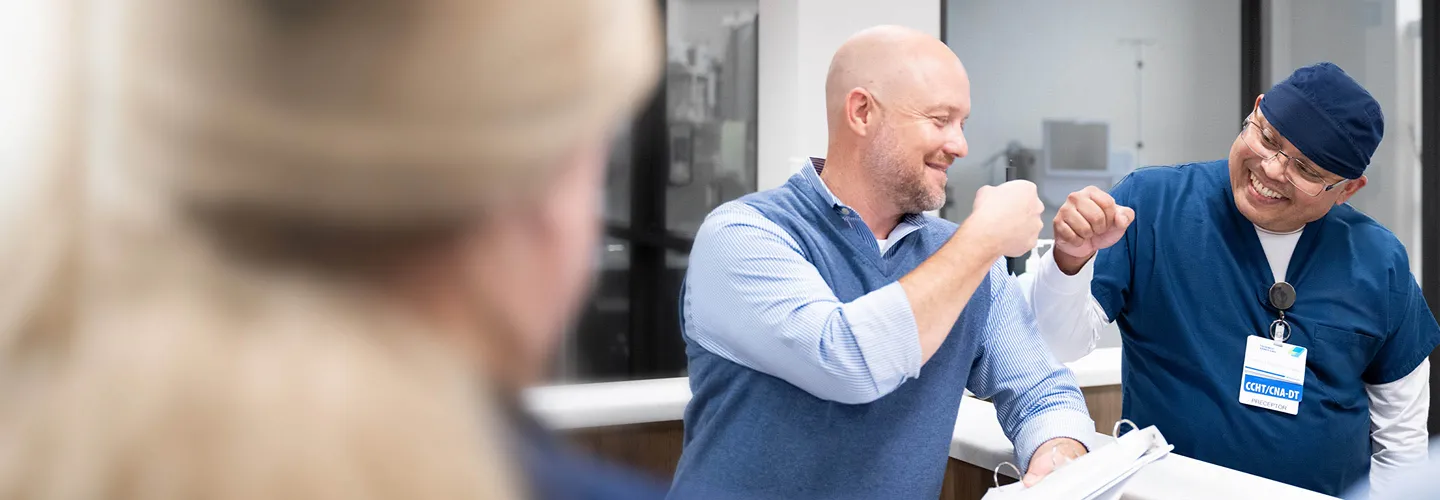 Two men smiling and giving each other a fist bump in a bright office setting; one wears business casual clothes, the other wears medical scrubs and a badge.