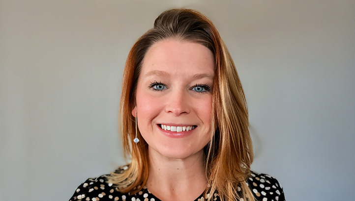 A woman with shoulder-length blonde hair, wearing a black and white polka dot top, smiles at the camera against a plain light gray background.