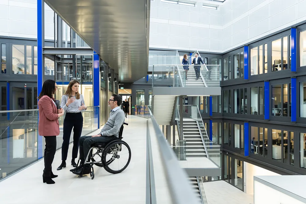 Three people, including a man in a wheelchair, have a conversation in a modern office corridor. Two more people are walking up a nearby staircase. The building features glass walls and an open, bright design.