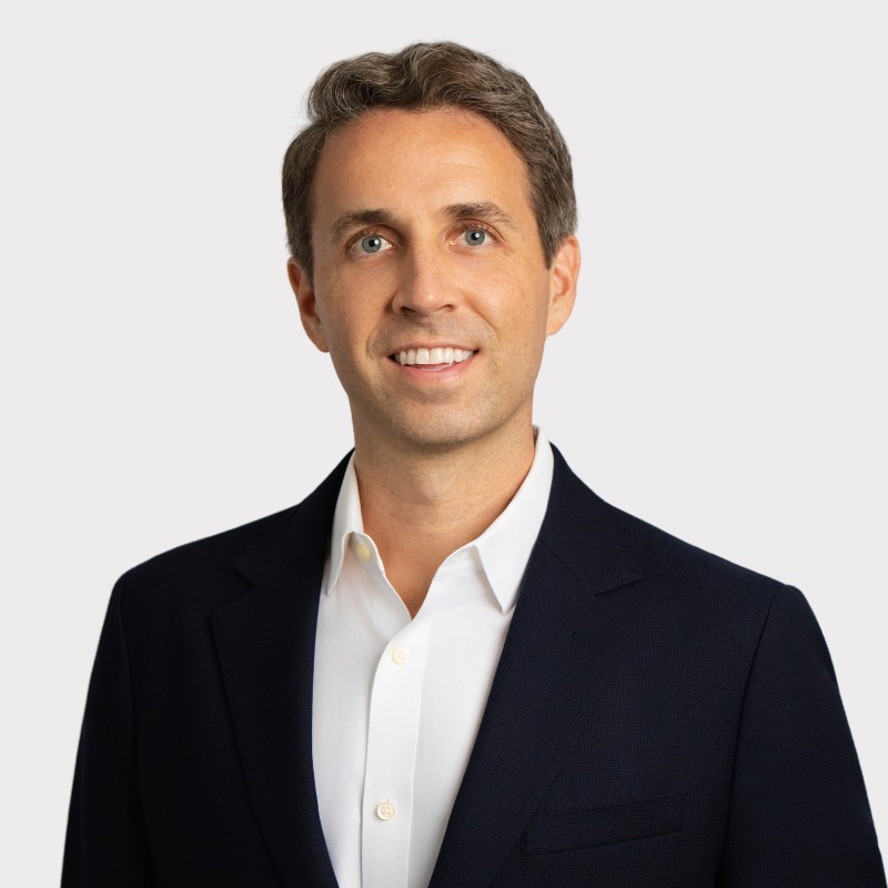 A man with short brown hair is smiling, wearing a white collared shirt and a dark suit jacket, standing in front of a plain light background.