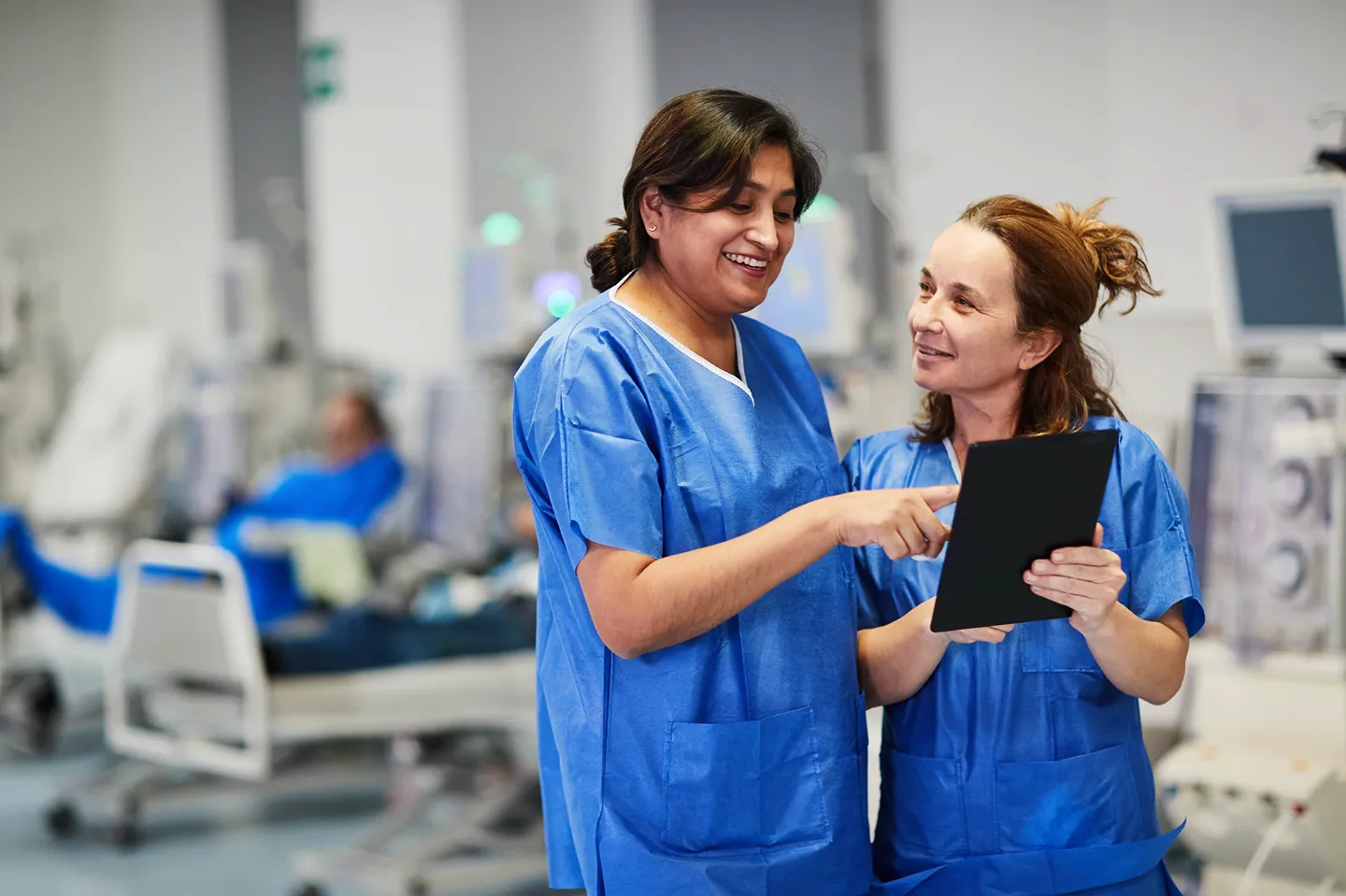 Two healthcare professionals in blue scrubs stand together, smiling and looking at a tablet device. One is pointing at the screen while the other holds the tablet. Medical equipment is visible in the blurred background.