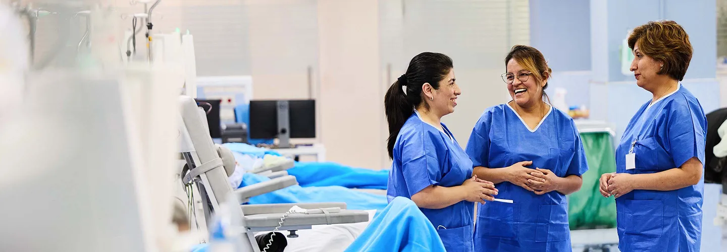 Three women in blue medical scrubs stand together in a brightly lit healthcare facility, smiling and talking. Medical equipment and hospital beds are visible in the background.