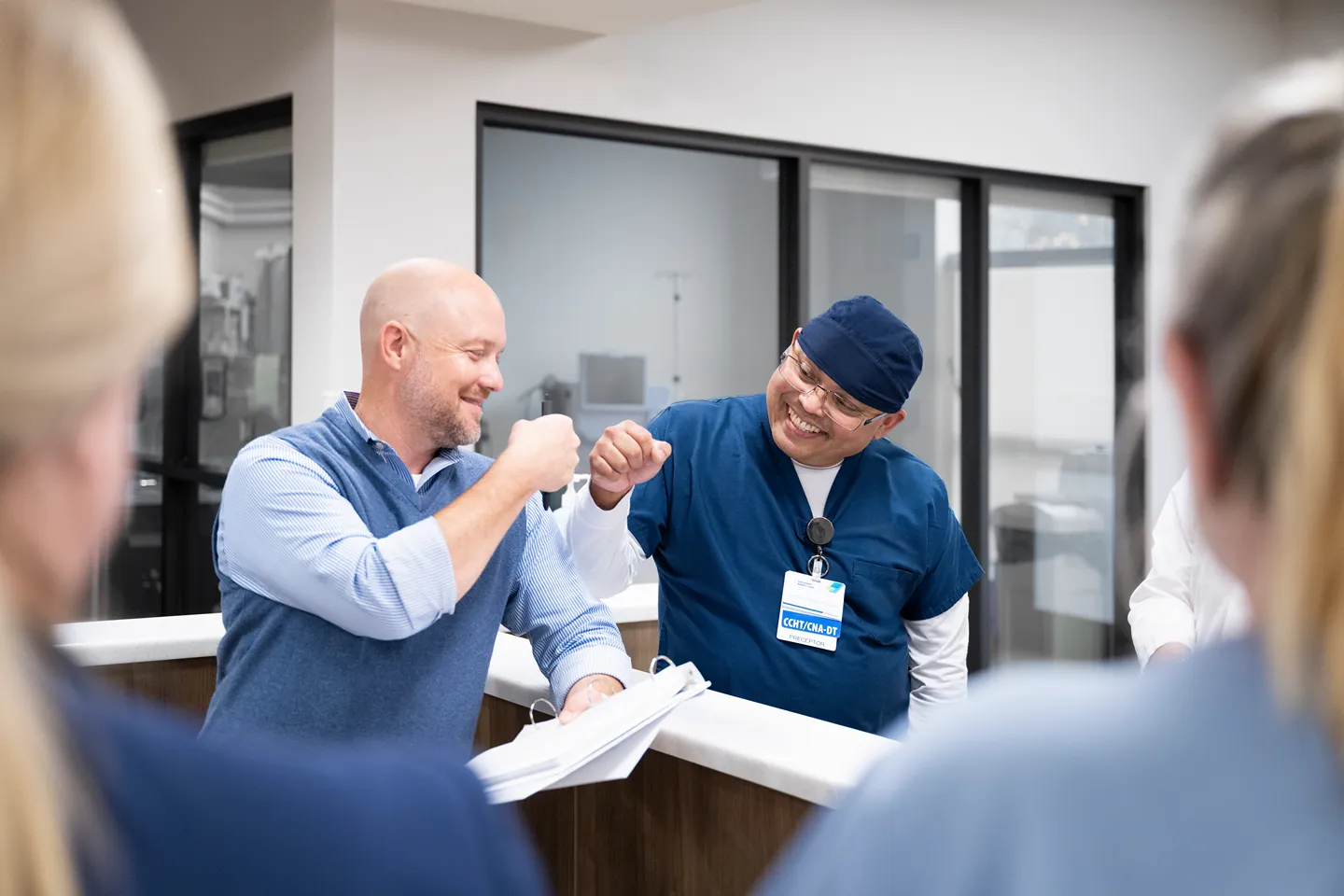 Two men, one in business casual clothes and the other in medical scrubs, smile and do a fist bump in an office or medical setting. One is holding a binder, and both appear to be celebrating or greeting each other.