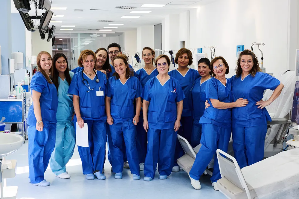 A group of healthcare professionals in blue scrubs stands together in a hospital corridor, smiling at the camera. Some have stethoscopes around their necks, and hospital beds are visible nearby.