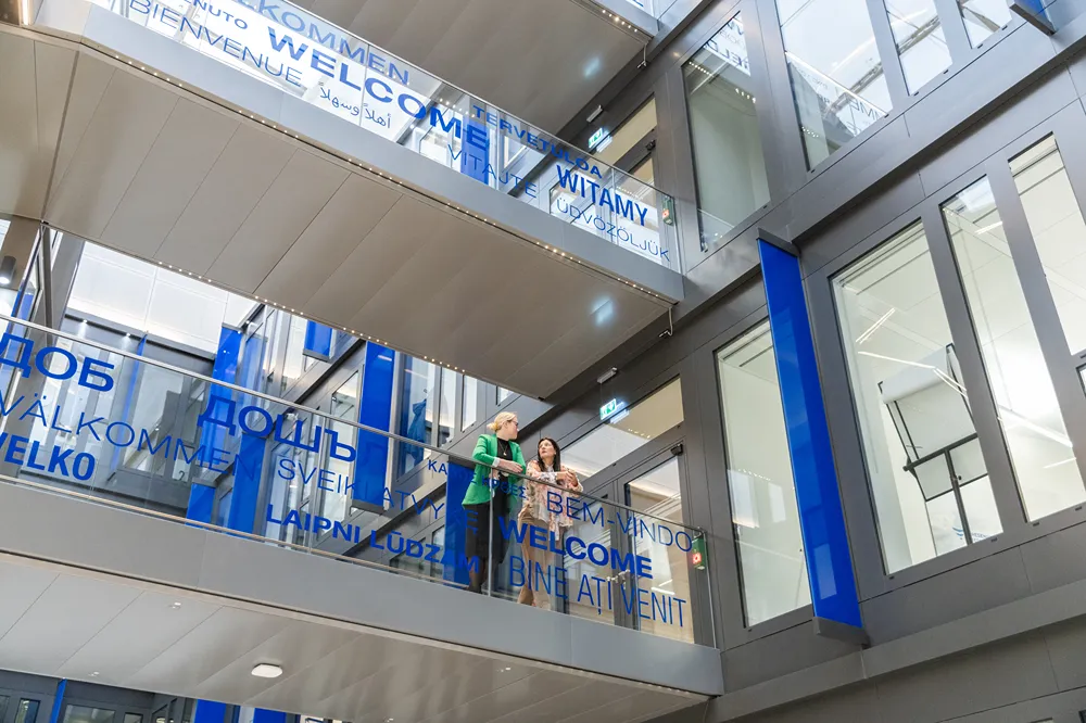 Two people stand and talk on an indoor balcony of a modern building decorated with the word 'Welcome' in multiple languages on glass panels. Blue and gray architecture is visible.