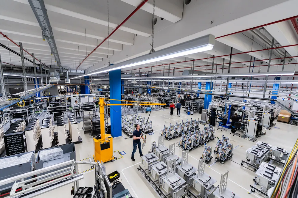 A spacious, brightly lit factory floor with workers operating machinery and assembling equipment. Organized rows of machines and tools are visible throughout the large industrial workspace.