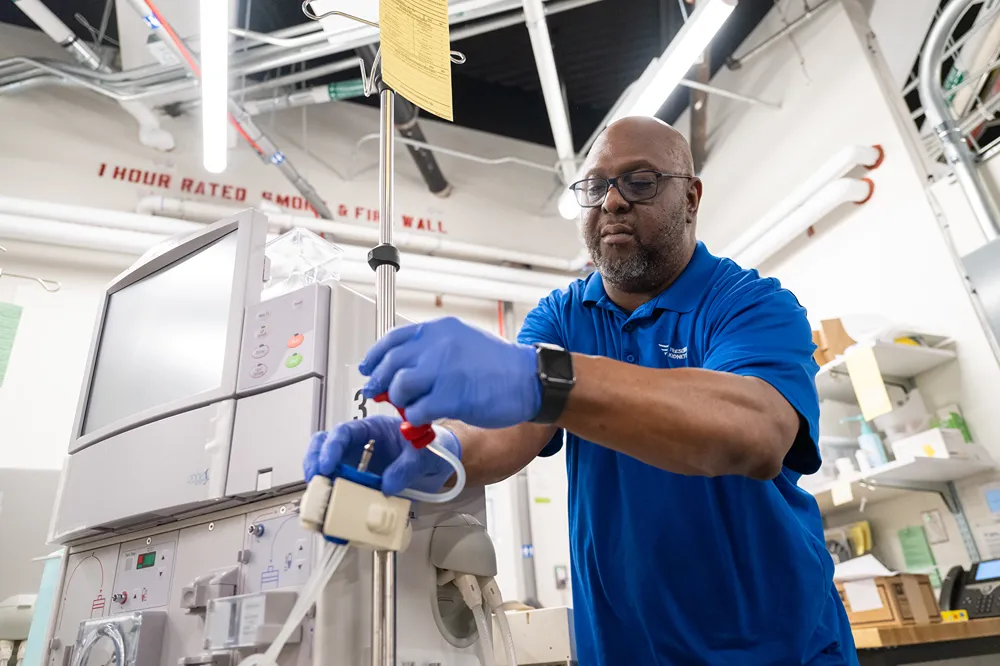 A man wearing blue gloves and a blue polo shirt adjusts medical equipment in a brightly lit industrial setting with pipes and machines in the background.