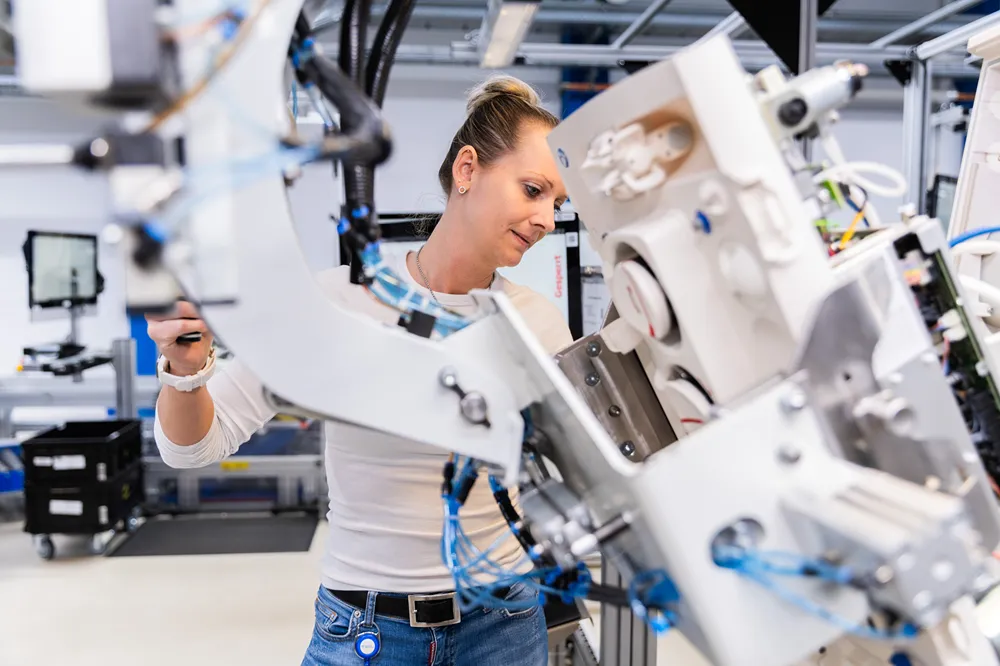 A woman in a white shirt operates and inspects a large, complex piece of machinery in a modern industrial or laboratory setting.