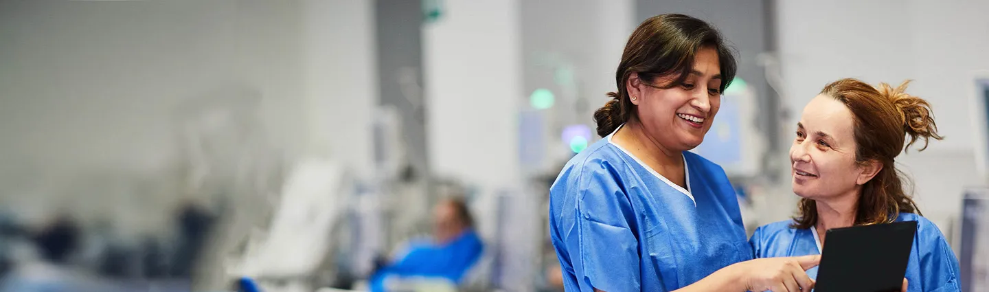 Two women in blue medical scrubs smile and look at a tablet together in a healthcare setting, with blurred medical equipment and personnel in the background.