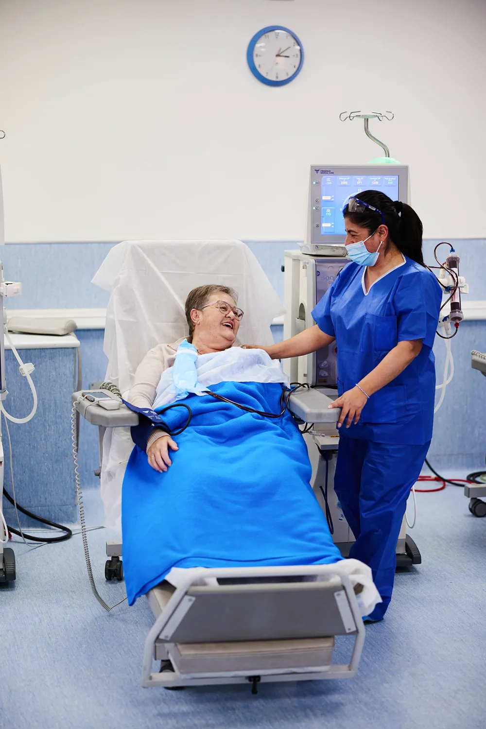 An older woman smiles while sitting in a hospital chair, receiving medical treatment. A nurse in blue scrubs and a face mask stands beside her, gently touching her arm and smiling. Medical equipment is visible in the background.