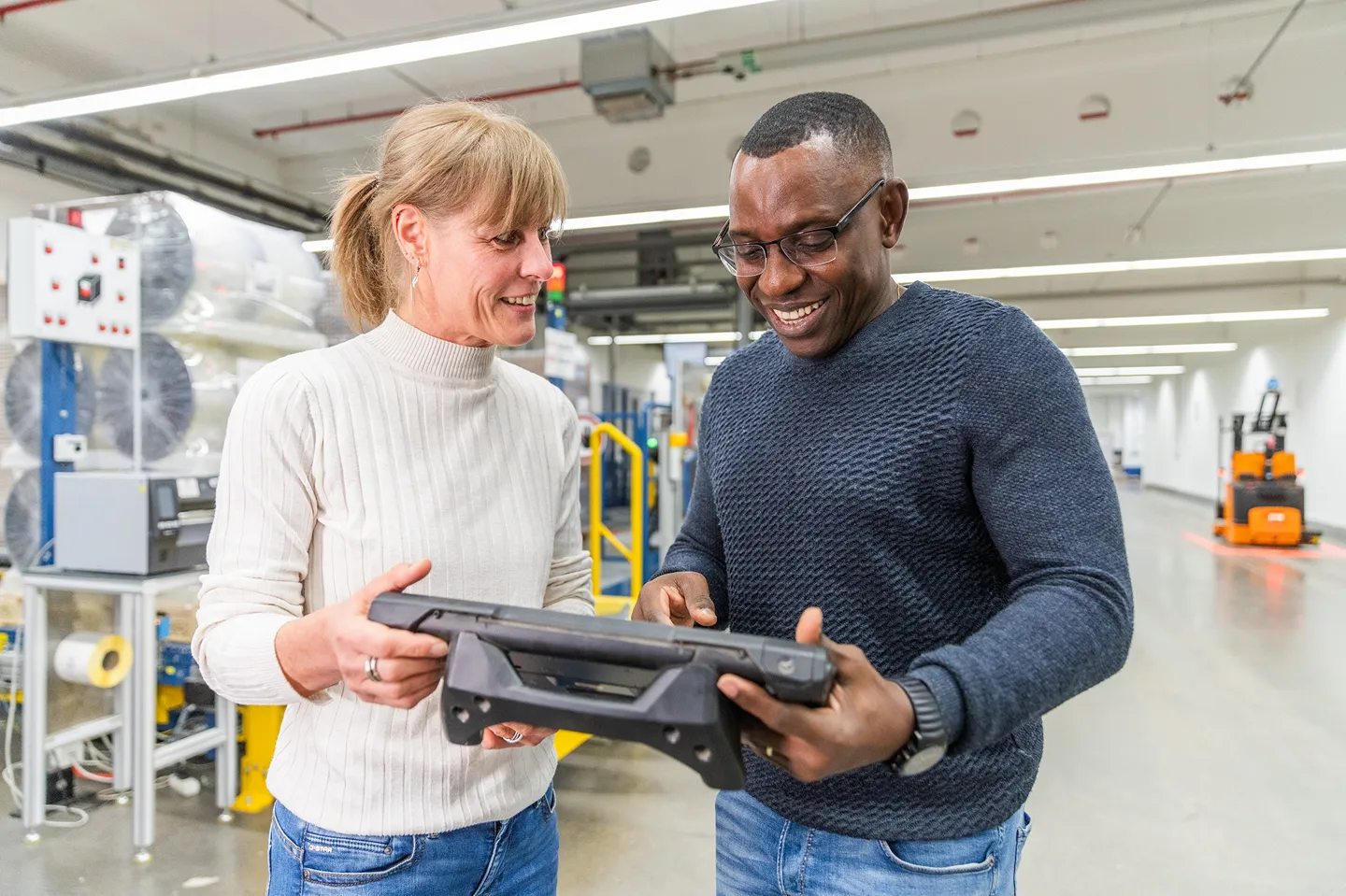 Two people standing in a bright industrial setting, smiling and looking at a tablet device together, with machinery and equipment visible in the background.