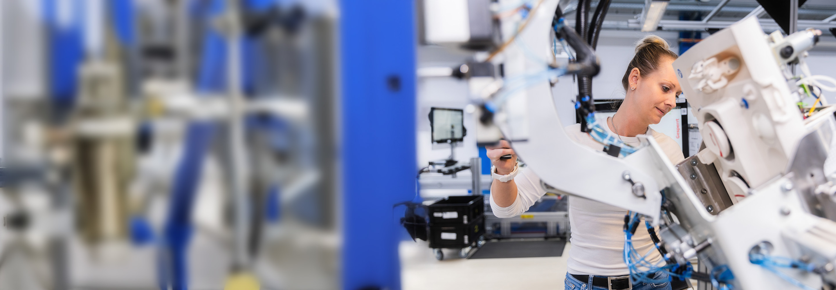 A woman in a white shirt works with advanced machinery in a modern, bright industrial or laboratory setting, focusing intently on the equipment.