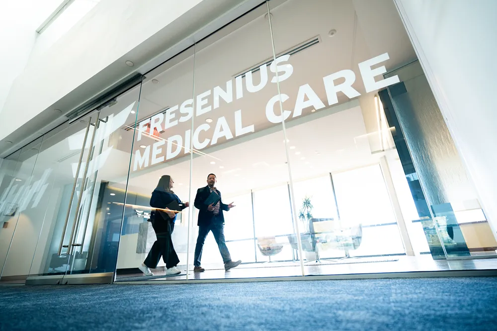 Two people walk through the glass entrance of a building with 'FRESENIUS MEDICAL CARE' written above the doors. The interior is bright with natural light and modern furnishings visible inside.