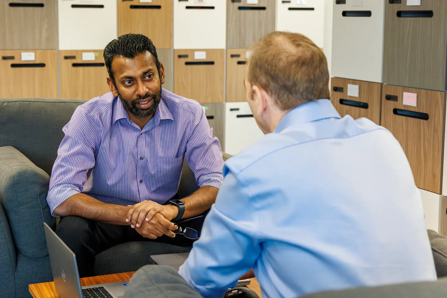 Two men sit facing each other and engage in conversation in an office setting, with lockers in the background. One man is holding eyeglasses while the other listens attentively.