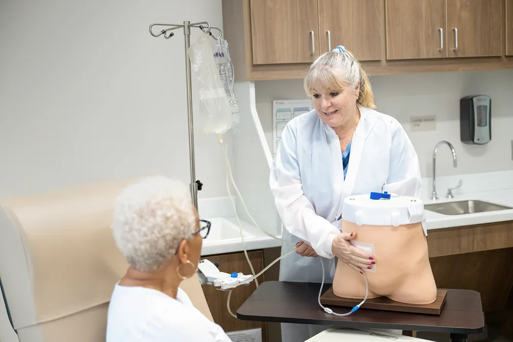 A healthcare professional demonstrates how to use a medical infusion device with a mannequin torso to an older woman seated nearby in a clinic setting.