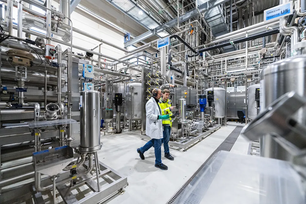Two people wearing safety gear walk through an industrial facility with stainless steel tanks, pipes, and machinery, surrounded by a clean, bright environment.