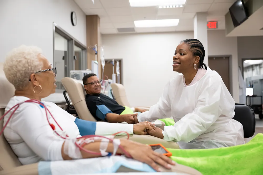A healthcare worker in protective clothing holds hands and smiles at an older patient receiving medical treatment, while another patient sits nearby in a clinic setting.