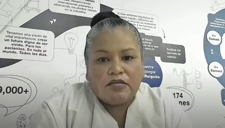 A woman with dark hair tied back, wearing a white top, sits in front of a wall covered with various charts, graphics, and text in Spanish.