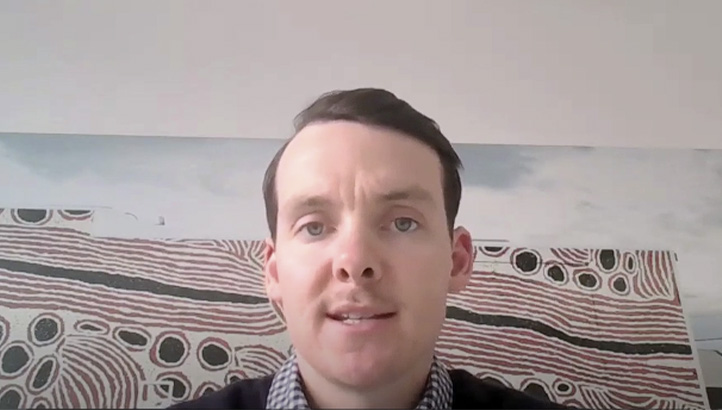 A man with short dark hair is speaking while facing the camera. He is indoors, in front of a wall with abstract lines and dot patterns in the background.