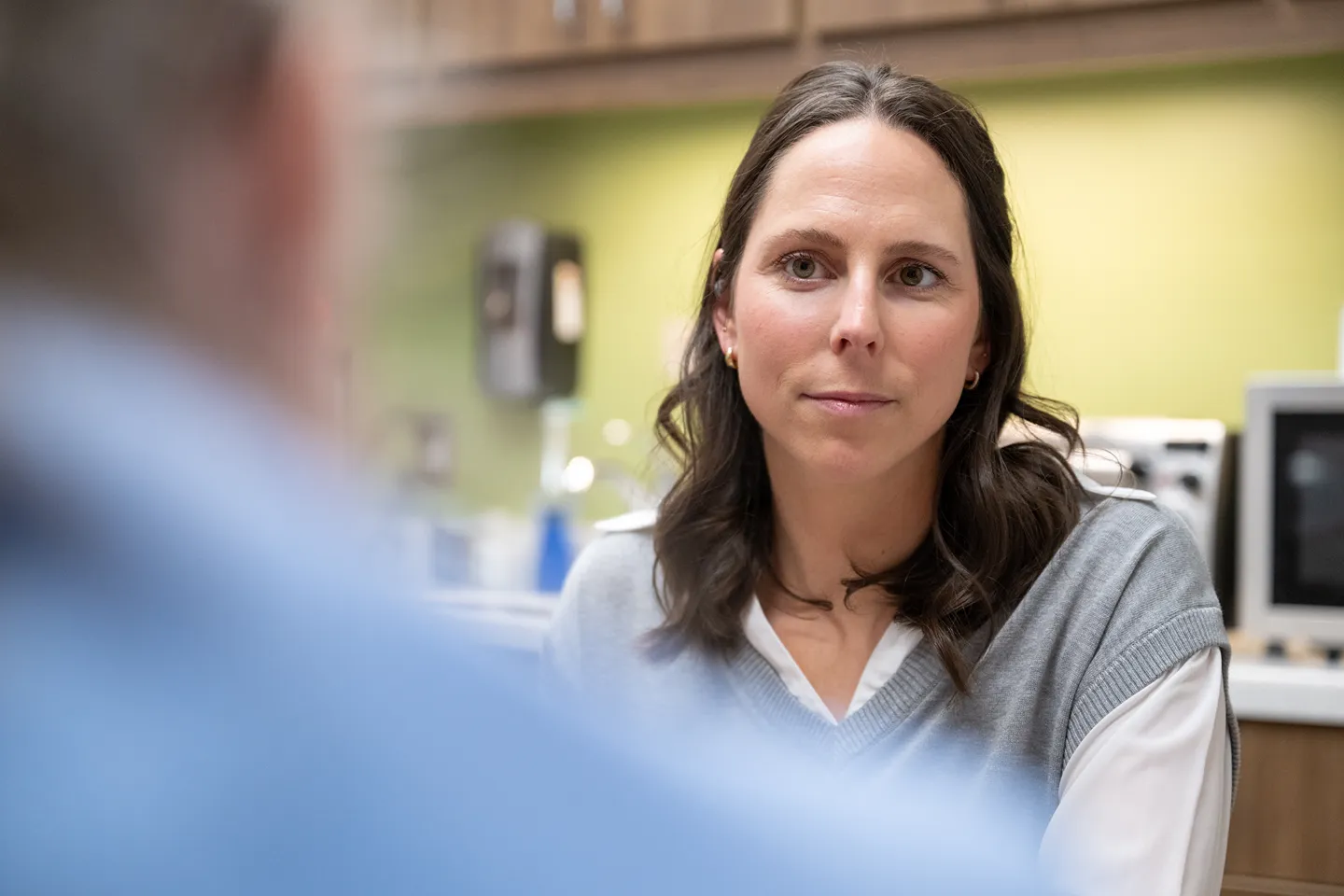 A woman with dark hair, wearing a gray vest over a white shirt, sits indoors and attentively listens to a person in the foreground, who is out of focus. The background is a softly lit kitchen or office space.