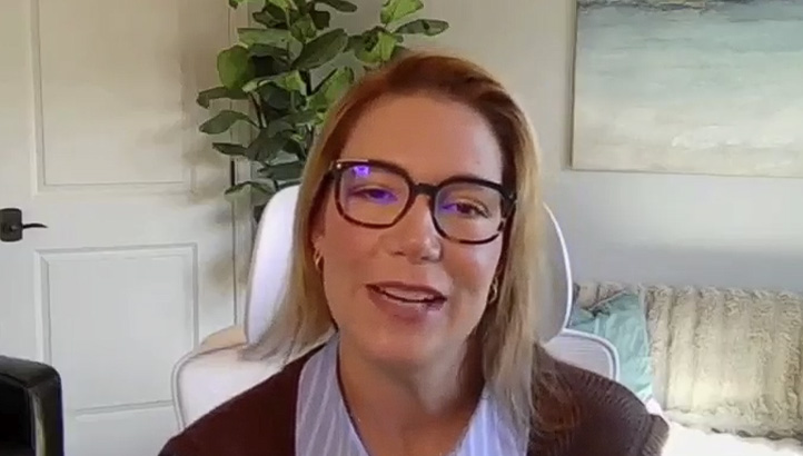 A woman with straight light brown hair and glasses smiles at the camera. She is wearing a light blue shirt and brown sweater, with a plant and painting visible in the background.