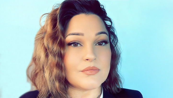 A woman with wavy, shoulder-length brown hair and natural makeup poses in front of a solid light blue background, wearing a dark blazer and white top.