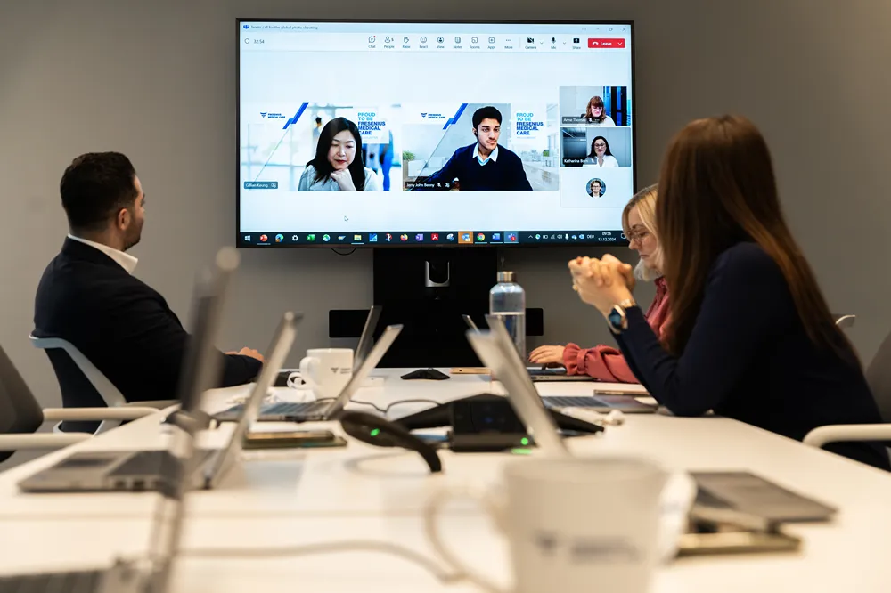 People sit around a table with laptops, participating in a video conference. A large screen displays multiple participants in a virtual meeting. Notebooks, water, and devices are visible on the table.
