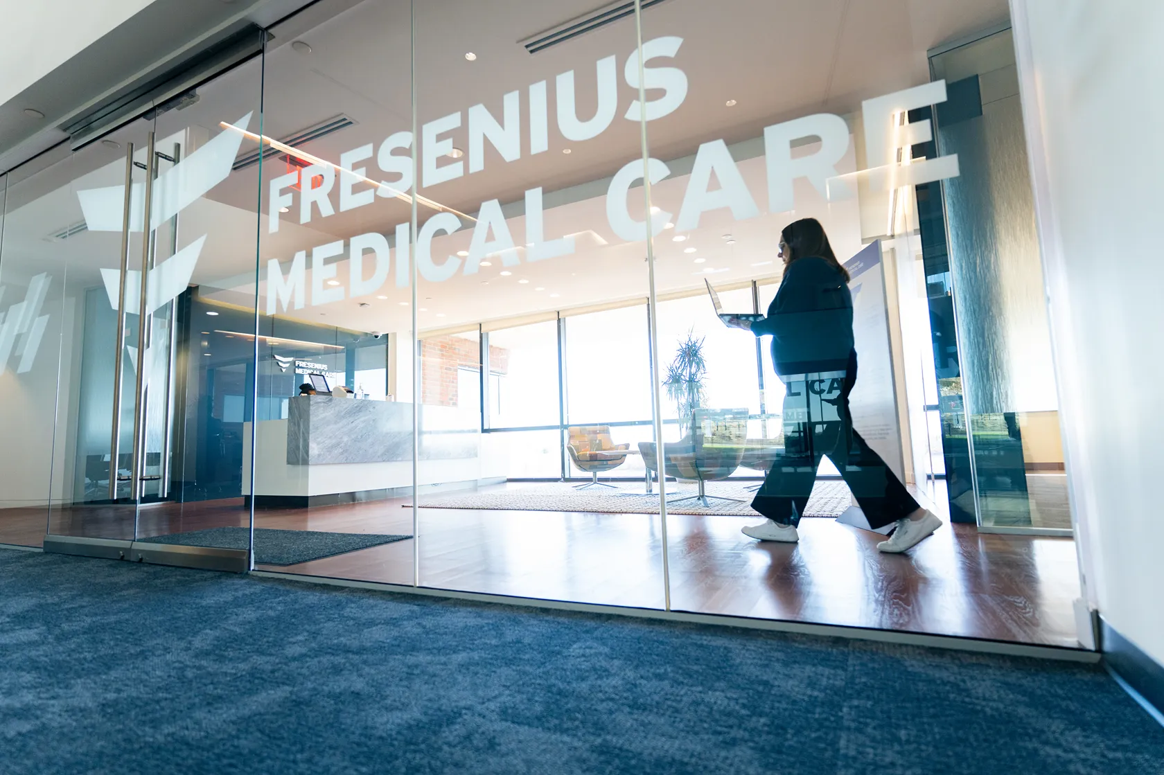A person holding a laptop stands inside a bright, modern office with large windows and frosted glass doors that have 'MEDICAL' written on them.