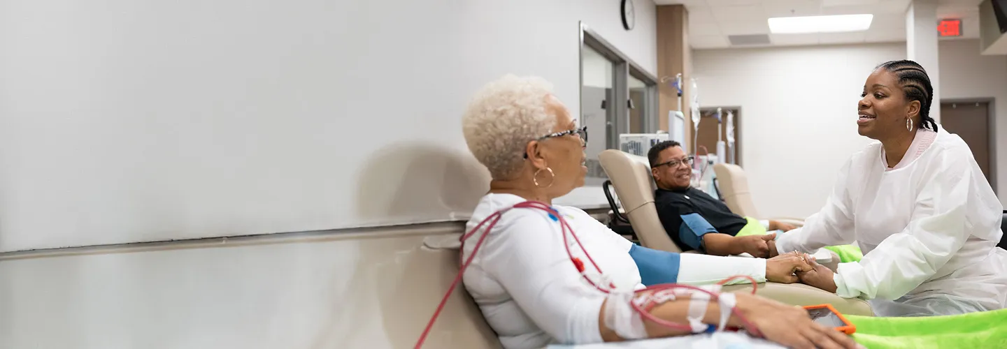 Two women sit in medical chairs, one receiving treatment with wires attached to her arm, while another woman in a white coat holds her hand and smiles; another patient sits in the background.