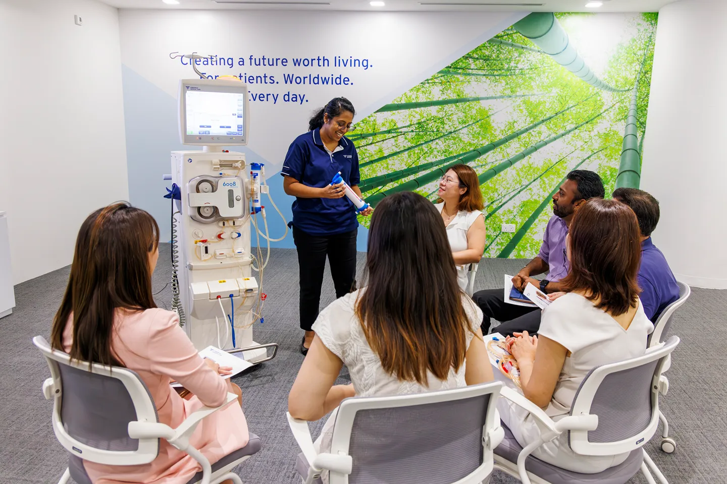 A woman in a blue shirt stands by a medical machine, speaking to a seated group of six adults in a bright room with a nature mural and the text “Creating a future worth living. For patients. Worldwide. Every day.”.