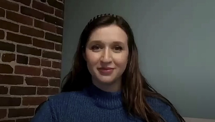 A woman with long, dark hair sits in front of a plain light-colored wall, wearing a gray hoodie and looking directly at the camera with a neutral expression.