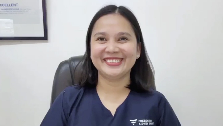 A woman with straight dark hair, wearing navy blue scrubs, sits smiling in an office chair against a light-colored wall with a framed certificate partially visible behind her.