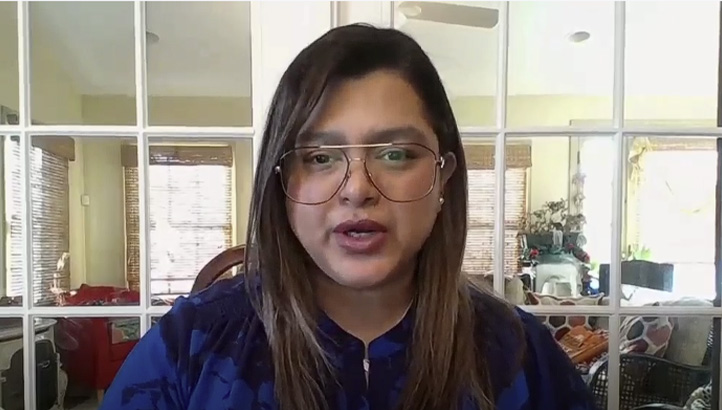 A person with long brown hair and glasses speaks while sitting indoors in front of glass doors, with various household items and light coming from windows in the background.