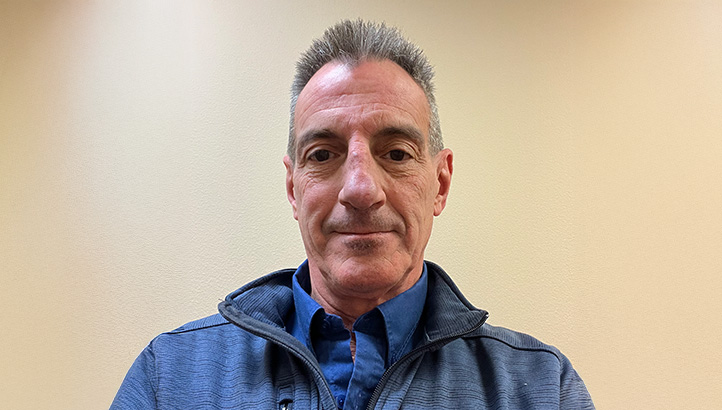 A middle-aged man with short gray hair wearing a blue collared shirt and a zip-up jacket, facing the camera against a plain beige background.