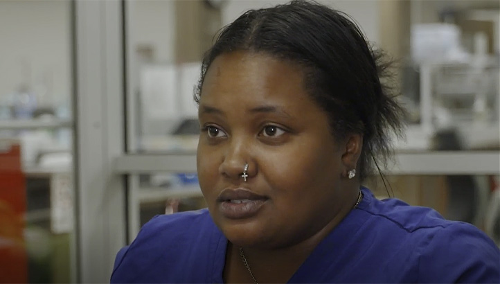 A woman with dark hair pulled back, wearing a blue top and a nose ring, looks slightly to the left. She is indoors, possibly in an office or medical setting, with blurred background details.