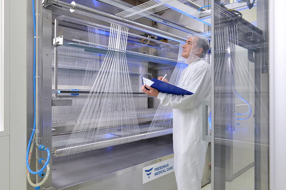 A person in protective clothing inspects a machine with multiple white threads at a Fresenius Medical Care facility, holding a clipboard and pen while observing the equipment.