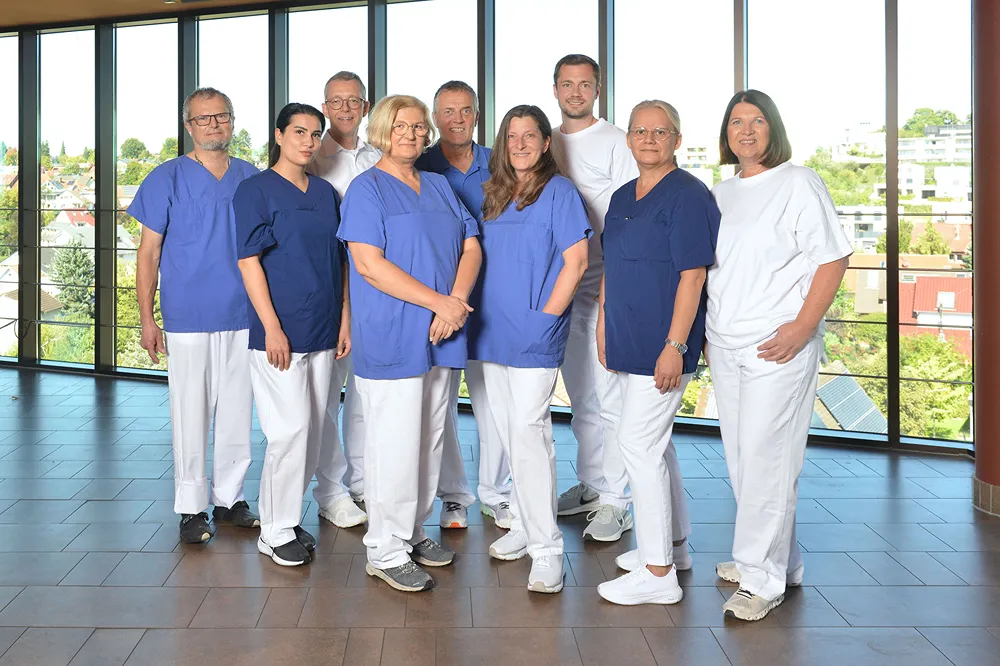A group of nine healthcare professionals, dressed in blue and white medical scrubs, stand together indoors in front of large windows with a view of greenery outside. They are smiling and posing for the photo.
